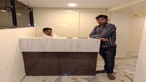 A reception area with a marble counter and a dark base, a person standing beside it, and another seated behind the counter. - Hotel Radhe (Vadodara)