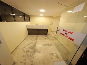 A narrow hallway with a reception desk, a sign reading "WELCOME TO HOTEL RADHE," and a tiled floor.