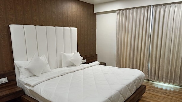 A modern hotel room with a large bed, wooden headboard, and a view of the city through sheer curtains. - Hotel Radhe (Vadodara)