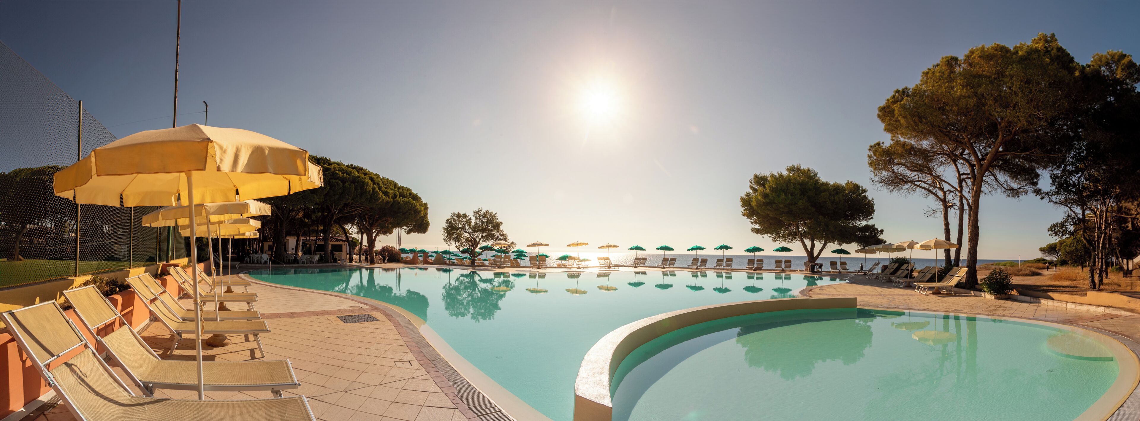 A swimming pool with lounge chairs and umbrellas, surrounded by trees and a clear sky.