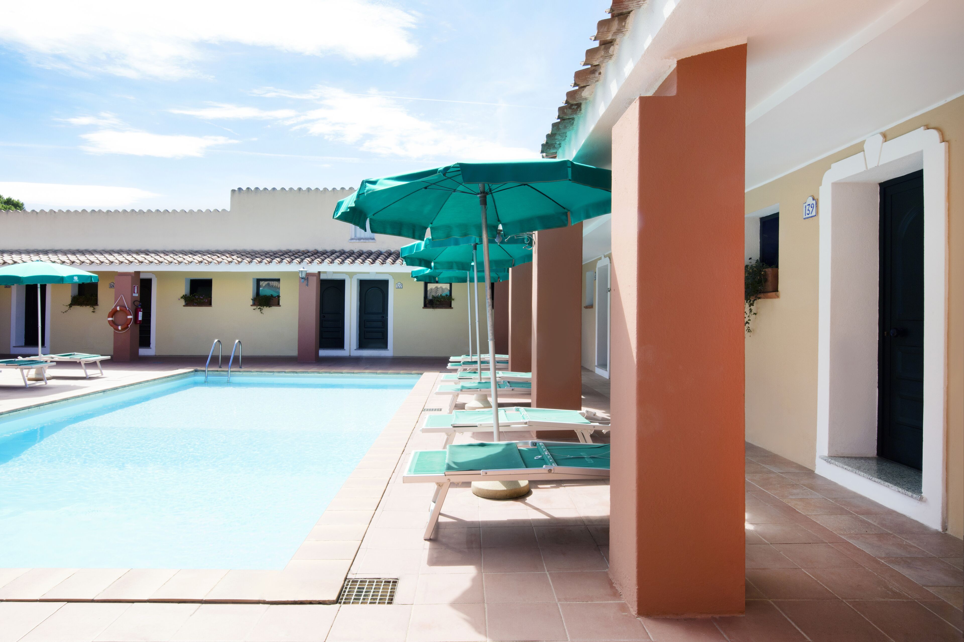 Ein Swimmingpool mit Liegestühlen und Sonnenschirmen, ein gefliestes Becken sowie ein Gebäude mit einem geziegelten Dach und beigen Wänden.