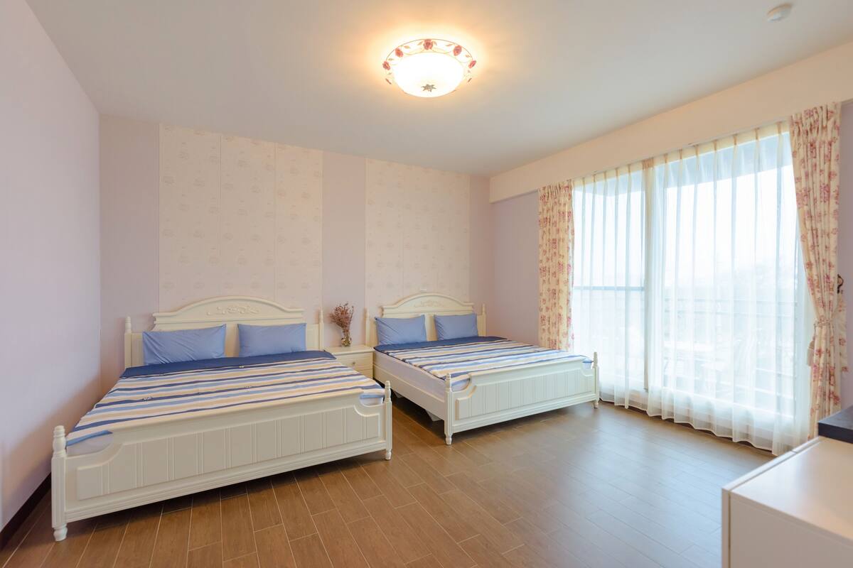 A bedroom with two beds, a nightstand, a lamp, and a window with curtains.