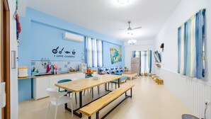 A modern kitchen with a long wooden table, white chairs, and a bench. There is a kitchen island with a sink and a microwave. The walls are painted light blue, and there is a ceiling fan.