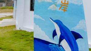 A surfboard with Chinese characters and a dolphin against a blue sky with clouds, placed in front of a white building with blue trim.