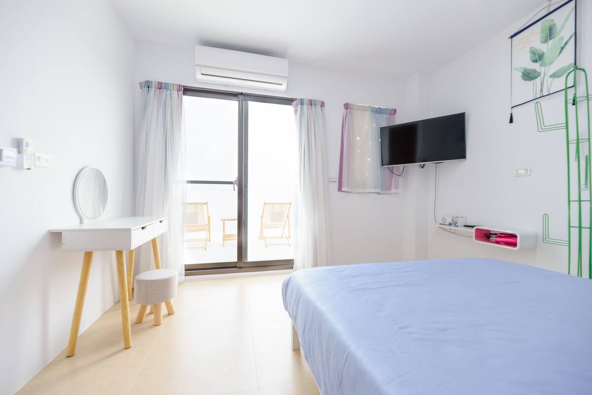 Habitación de hotel con cama, escritorio con espejo, televisor montado en la pared y puerta corrediza de vidrio que da a un balcón.