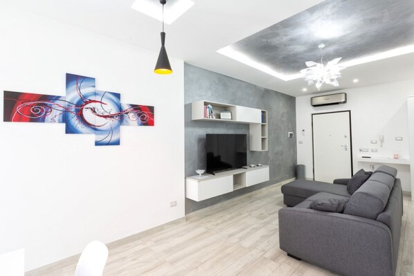 Un salon moderne avec un canapé gris, un téléviseur à écran plat, des étagères blanches intégrées et des œuvres d’art abstraites accrochées au mur.