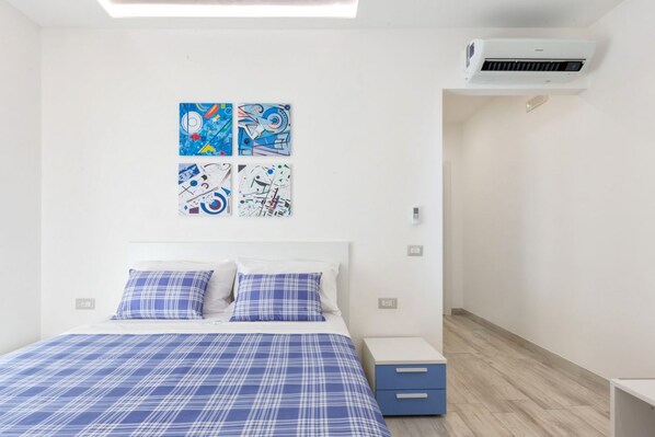 Une chambre à coucher comprenant un lit, deux oreillers, une table de chevet, un tiroir et quatre tableaux abstraits accrochés au mur.