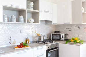 Une cuisine moderne avec des meubles blancs, une cuisini ère à gaz, un four à micro-ondes et une machine à café. On peut voir des bocaux sur les étagères et une planche à découper avec des légumes sur le plan de travail.