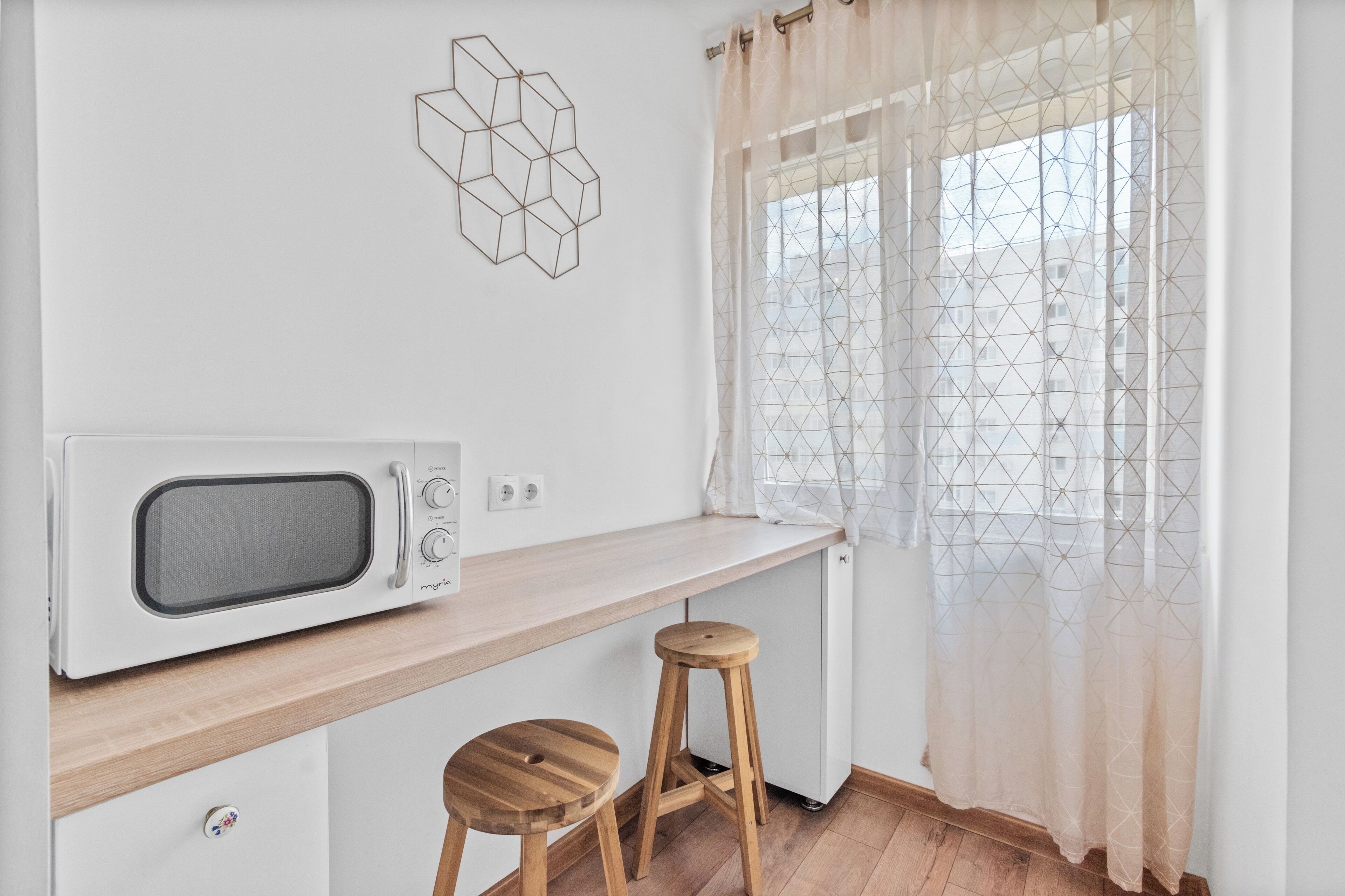 Une petite cuisine équipée d’un four à micro-ondes, de tabourets en bois et d’une fenêtre agrémentée de rideaux à motifs géométriques.