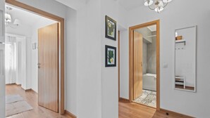 Un couloir doté d’un plancher en bois, de murs blancs et d’une porte donnant sur une salle de bain. Des tableaux encadrés ornent le mur, et un miroir est placé au-dessus de la porte.