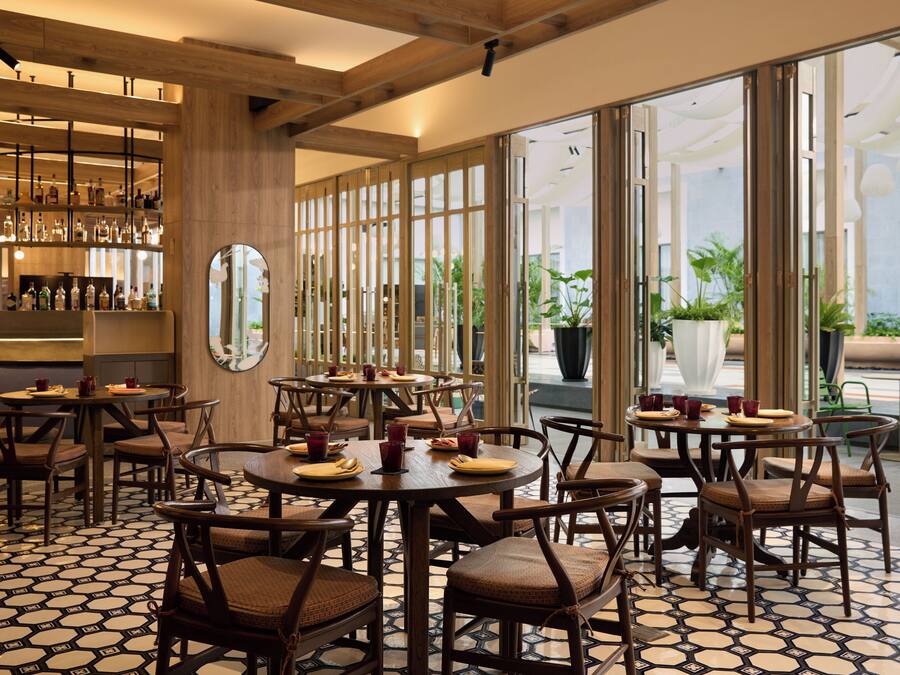 A restaurant with round tables and chairs, a bar area with bottles, and large windows with plants outside.