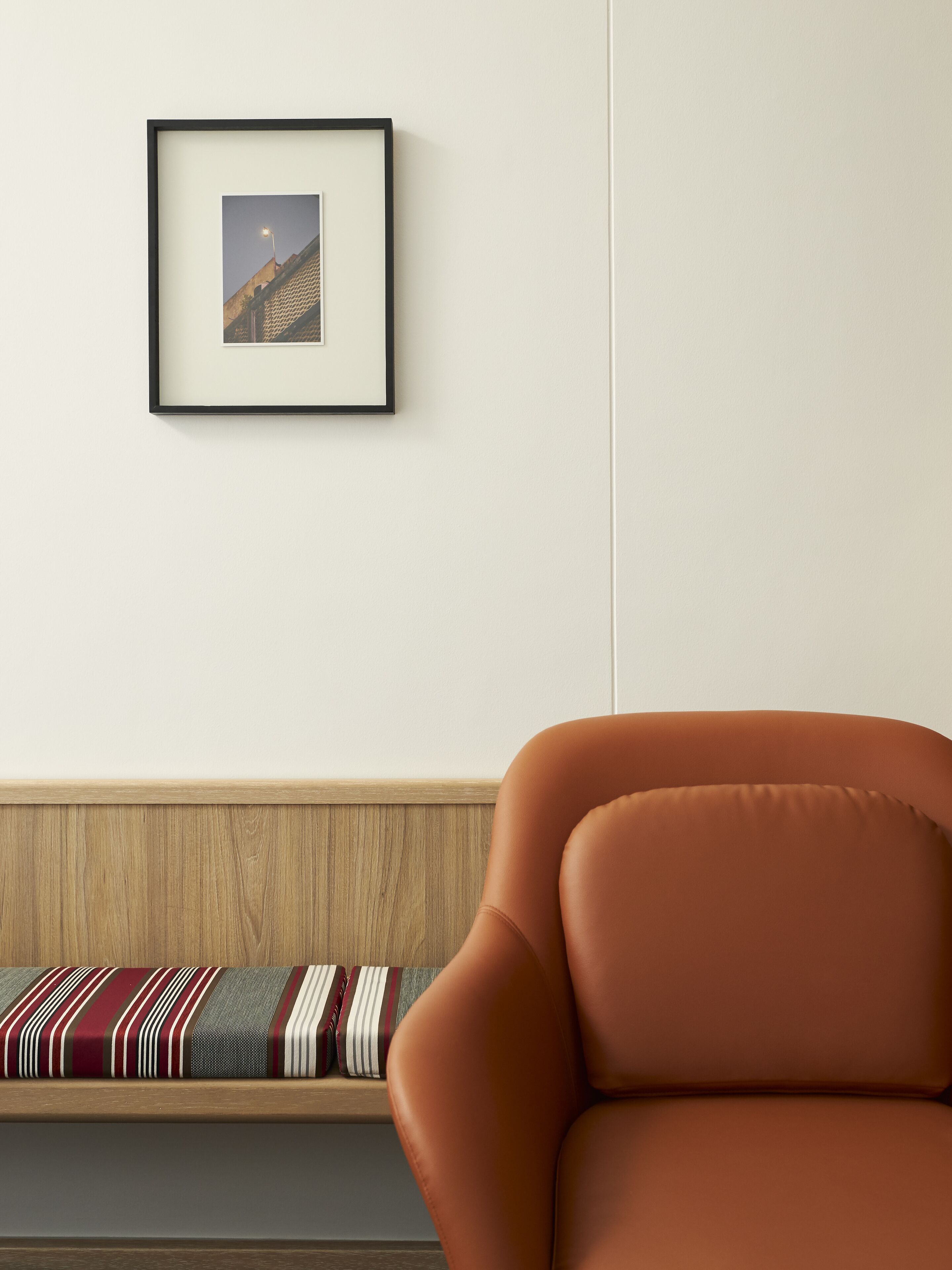 A room with a framed picture on the wall, a wooden bench with a striped cushion, and an orange chair.