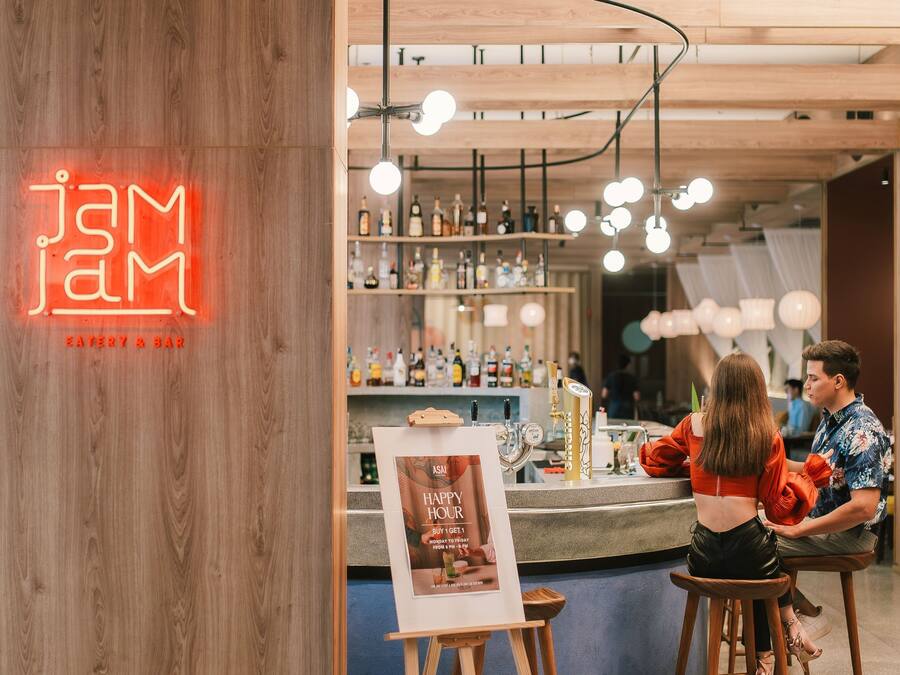 Un bar avec une enseigne au néon, des étagères en bois remplies de bouteilles et une affiche accrochée à un tabouret.