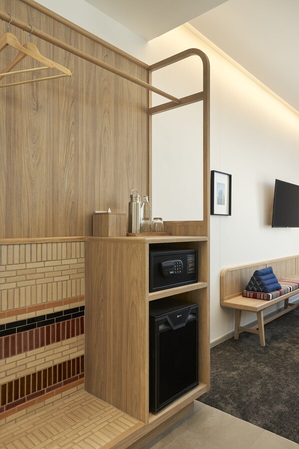 A hotel room with a wooden wardrobe, a black minibar, a wooden bench, and a television mounted on the wall.
