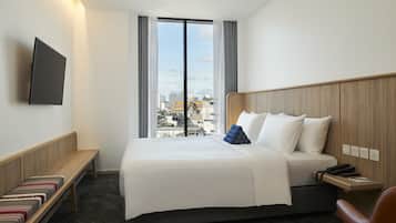 Habitación de hotel con una cama grande, un banco, un televisor y vistas a la ciudad a través del ventanal.
