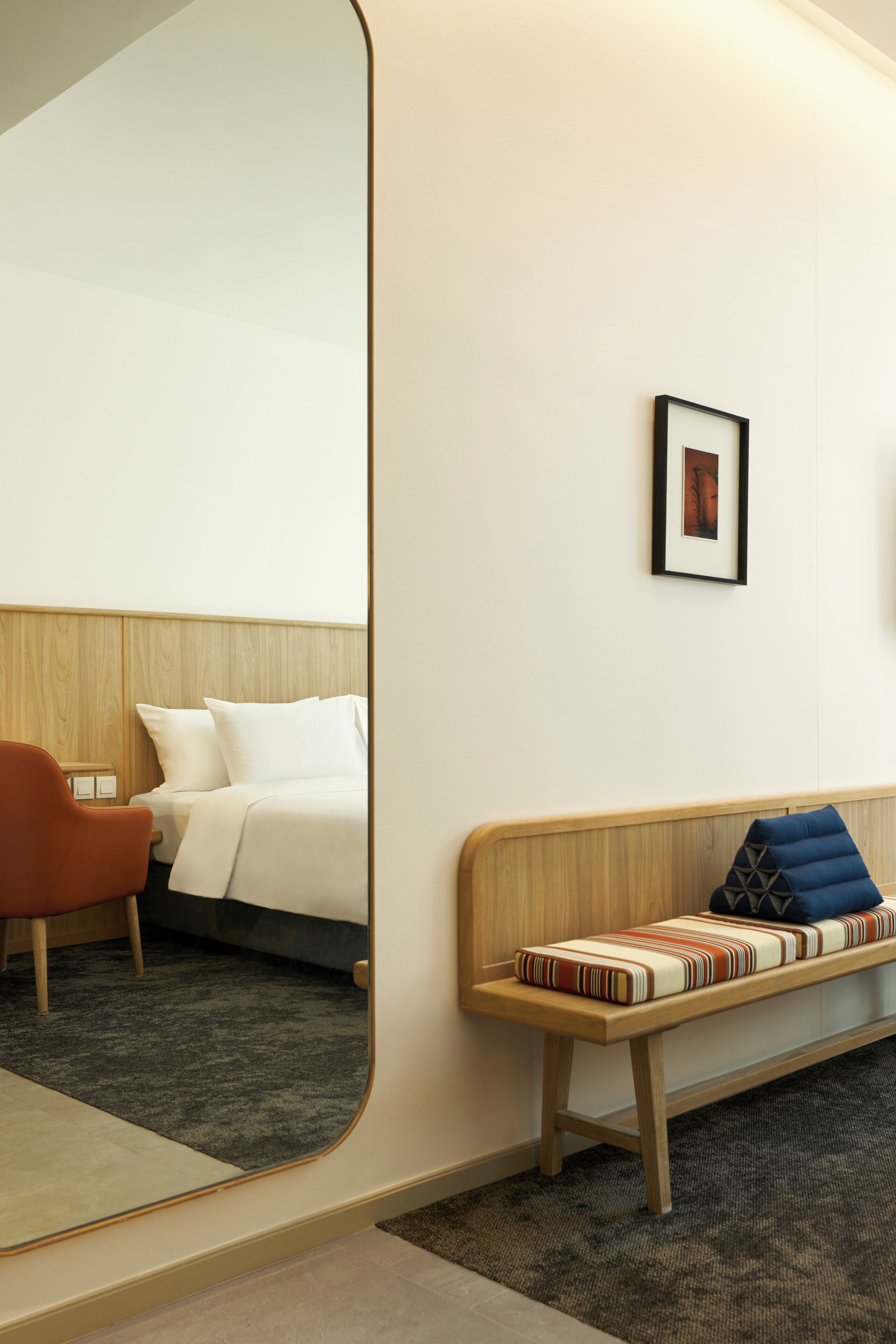 A hotel room with a bed, a chair, a bench, a framed picture on the wall, and a mirror reflecting part of the room.