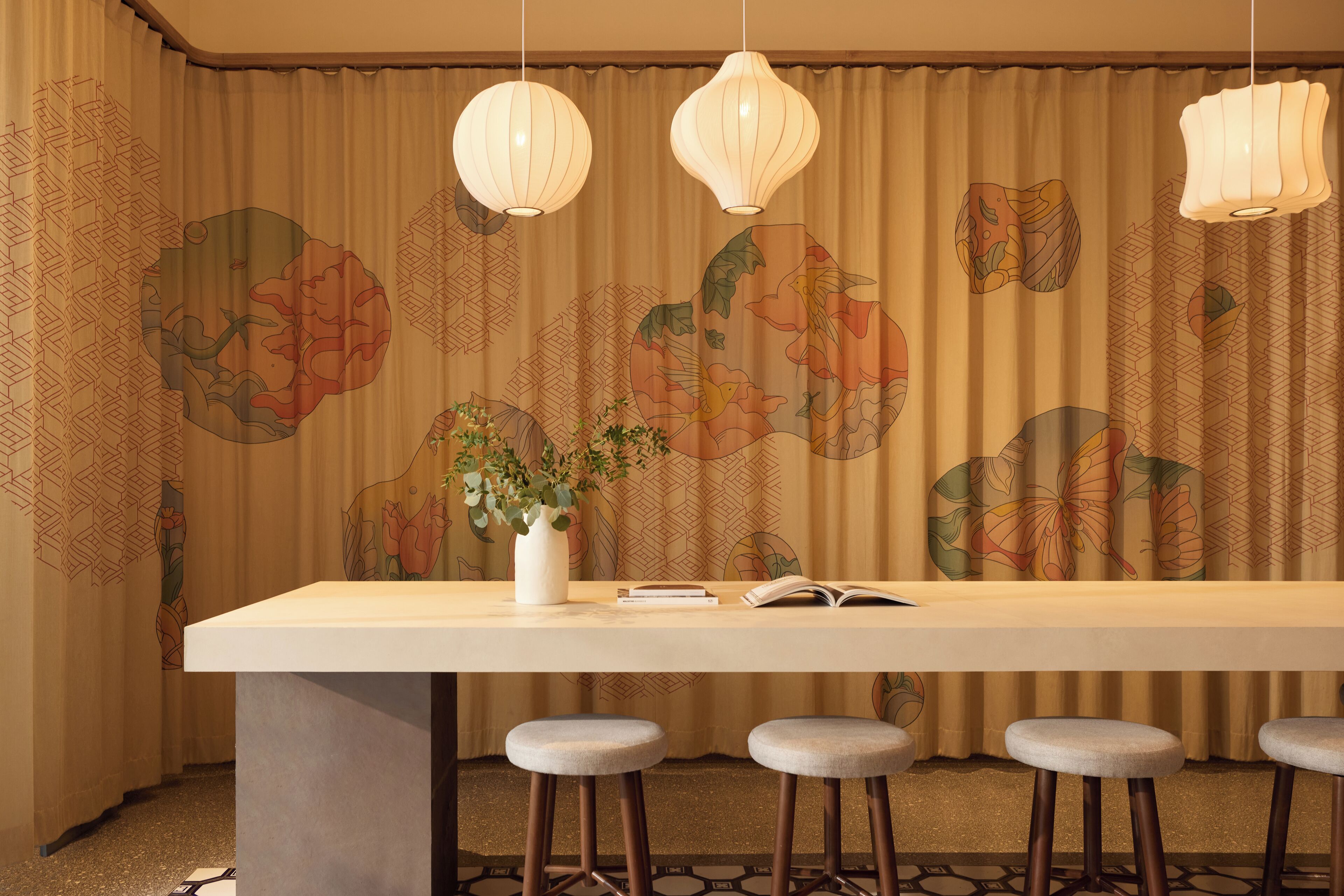 Un comedor con una mesa larga, dos taburetes de bar, un florero con flores y lámparas colgantes.