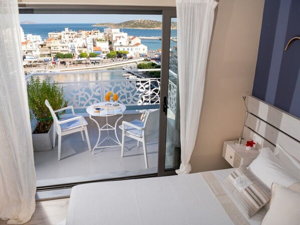 A balcony with a view of a coastal town, a small table with two chairs, and a bed with white bedding.