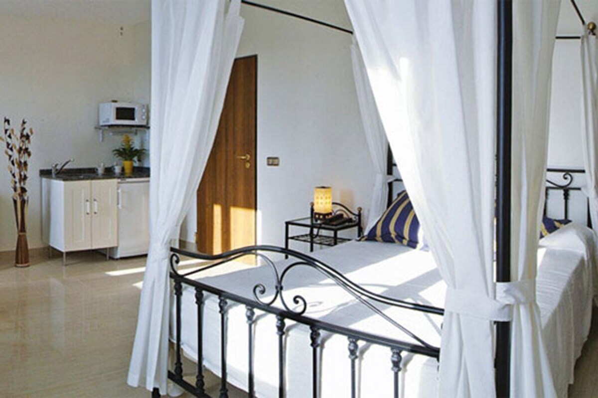 A bedroom with a four-poster bed, a nightstand, a lamp, a chair, a small table, and a kitchenette in the background.
