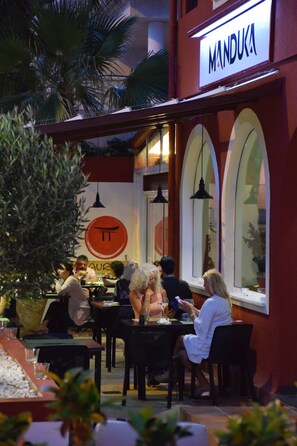 Un área de comedor al aire libre con mesas y sillas, palmeras y un letrero que dice "MANDUKA".