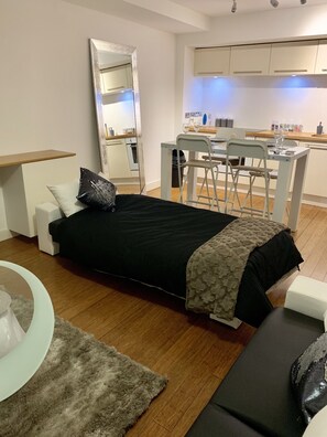 A modern bedroom with a bed, a small desk, a chair, and a kitchenette in the background.