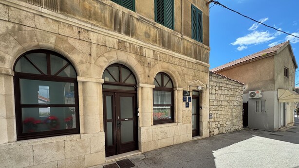 Un edificio de piedra con ventanas arqueadas y una puerta, un pequeño letrero sobre la puerta, y un cuadro eléctrico en la acera.