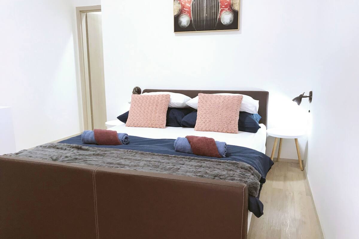 A neatly made bed with a brown headboard, a white bedside table, and a wall-mounted lamp.