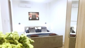 Un dormitorio con cama, almohadas, mesita de noche, aire acondicionado de pared y un cuadro en la pared.