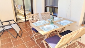 Une table de salle à manger en bois, dressée pour quatre personnes, avec des sets de table bleus et des assiettes blanches, une bouteille de vin et des verres, le tout sur une terrasse carrelée.