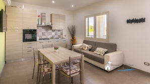 A modern kitchen with wooden cabinets, a dining table with chairs, and a sofa in a living area with a large window.
