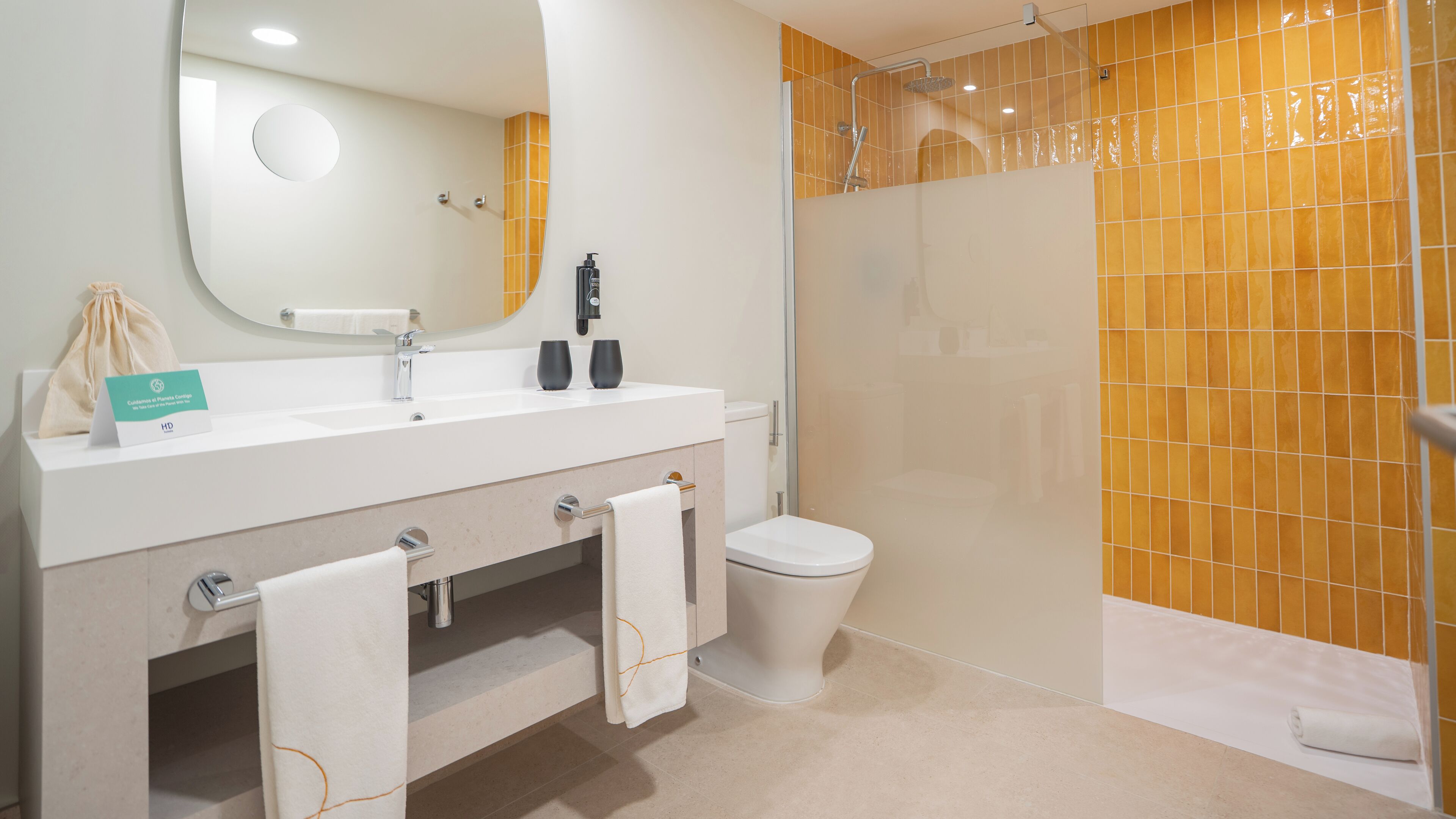 Een moderne badkamer met een wit wastafelmeubel, spiegel en toilet, een douche met gele tegels, en een handdoekrek.