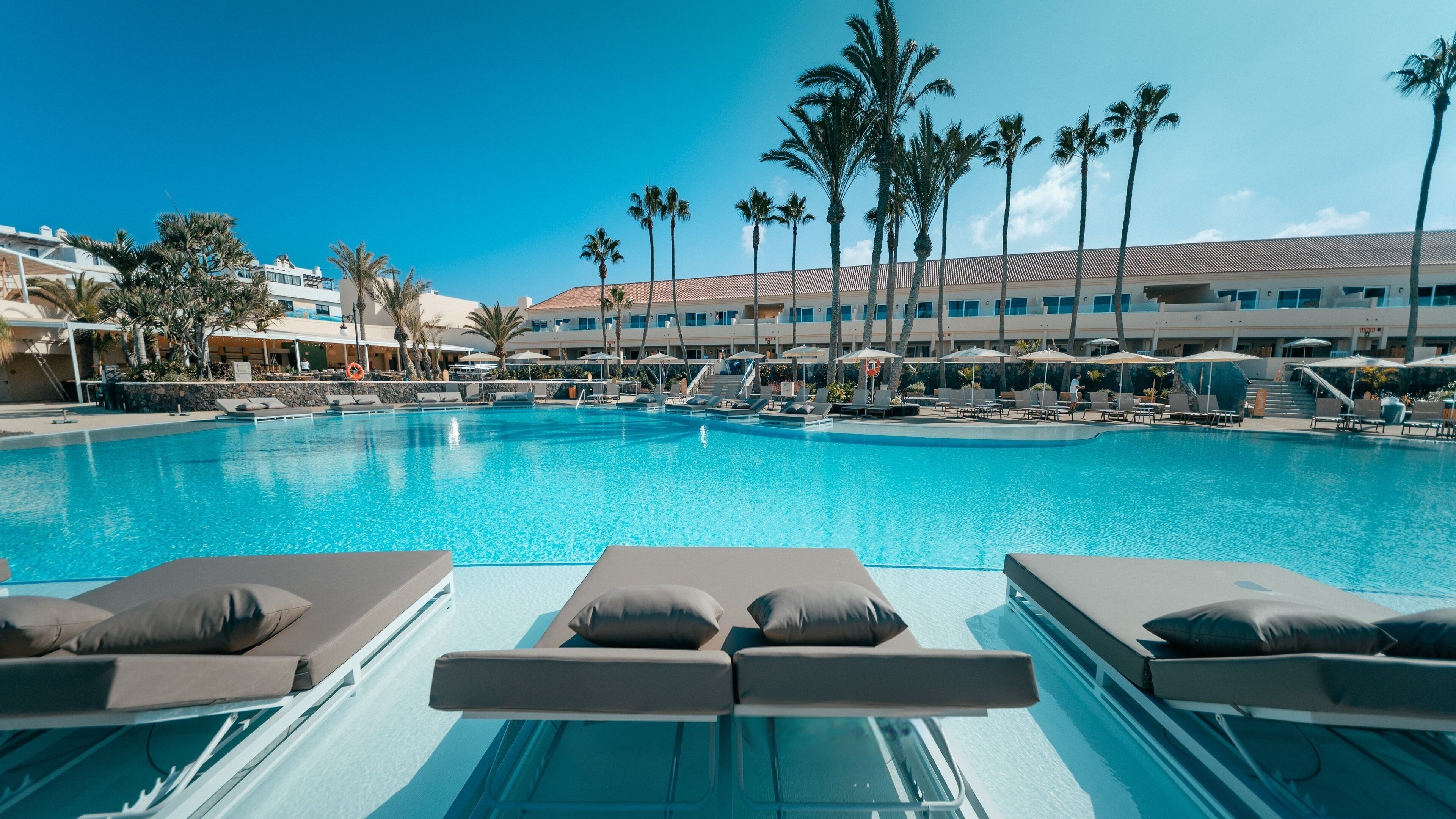 Una gran piscina con sillas de descanso y sombrillas, rodeada de palmeras y edificios de hotel.