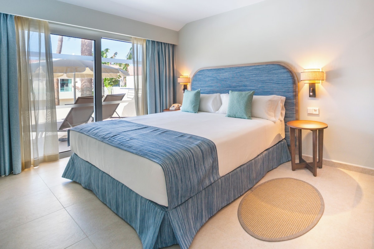 A hotel room with a large bed, a small table, a chair, and a view of the outdoors.
