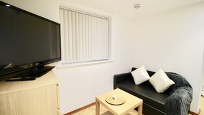 Ein Wohnzimmer mit einem Flachbildfernseher auf einer Holzkommode, einem schwarzen Sofa mit weißen Kissen und einem kleinen Holztisch mit einer Dekoration.