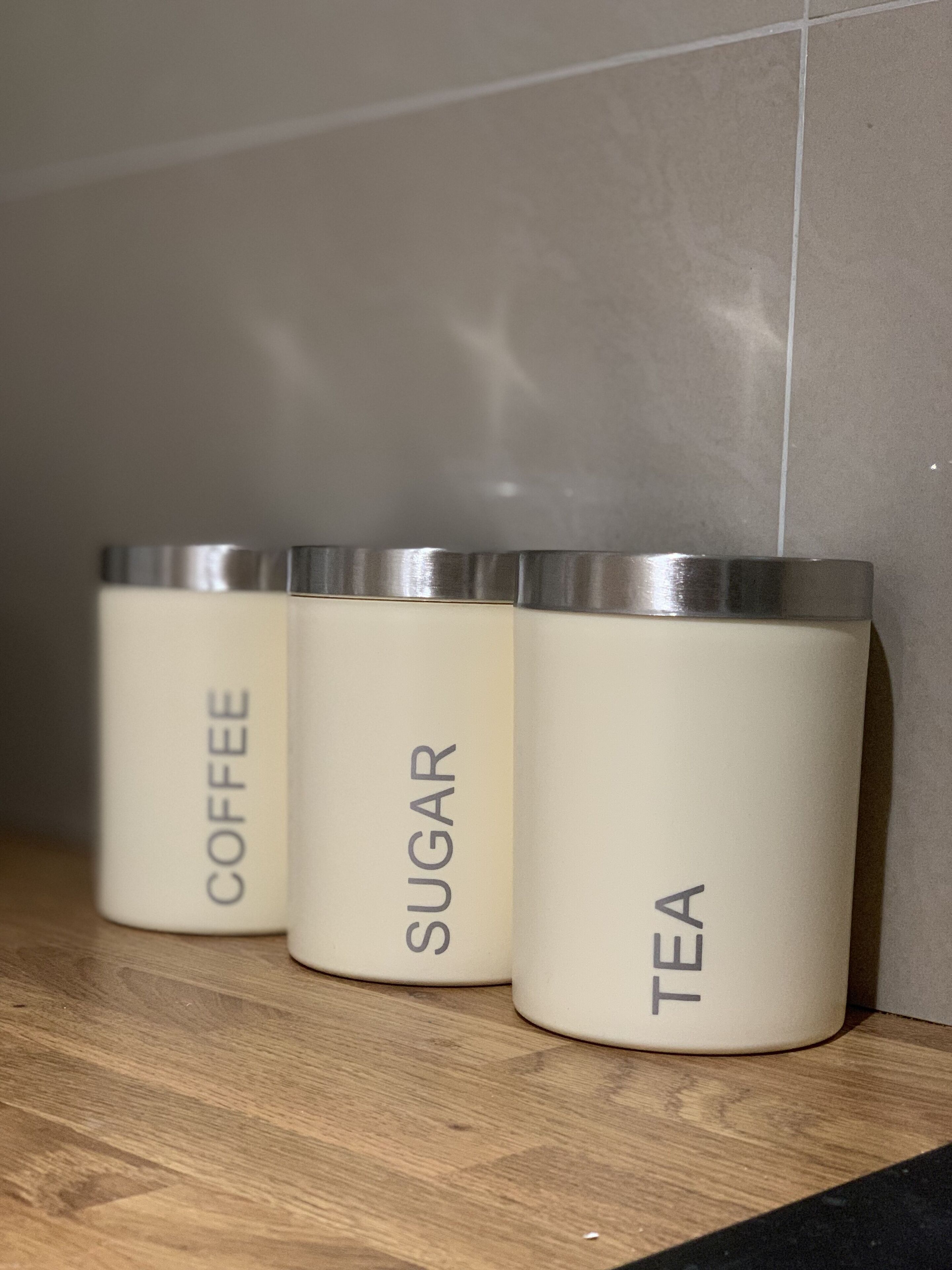 Drie witte bakken met zilveren deksels, gelabeld met 'COFFEE', 'SUGAR' en 'TEA', staan op een houten plank.