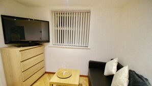 Ein kleines Wohnzimmer mit Fernseher auf einer Holzkommode, einem kleinen Tisch mit Dekoration und einem schwarzen Sofa mit Kissen.