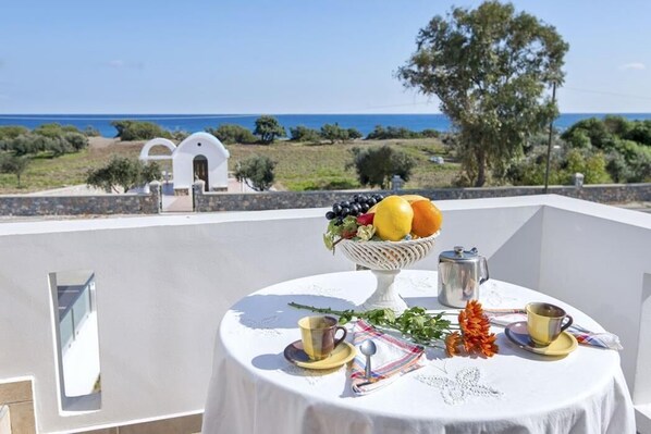 Una mesa dispuesta con fruteros, cafetera y tazas en un balcón con vista al mar y un edificio blanco al fondo.