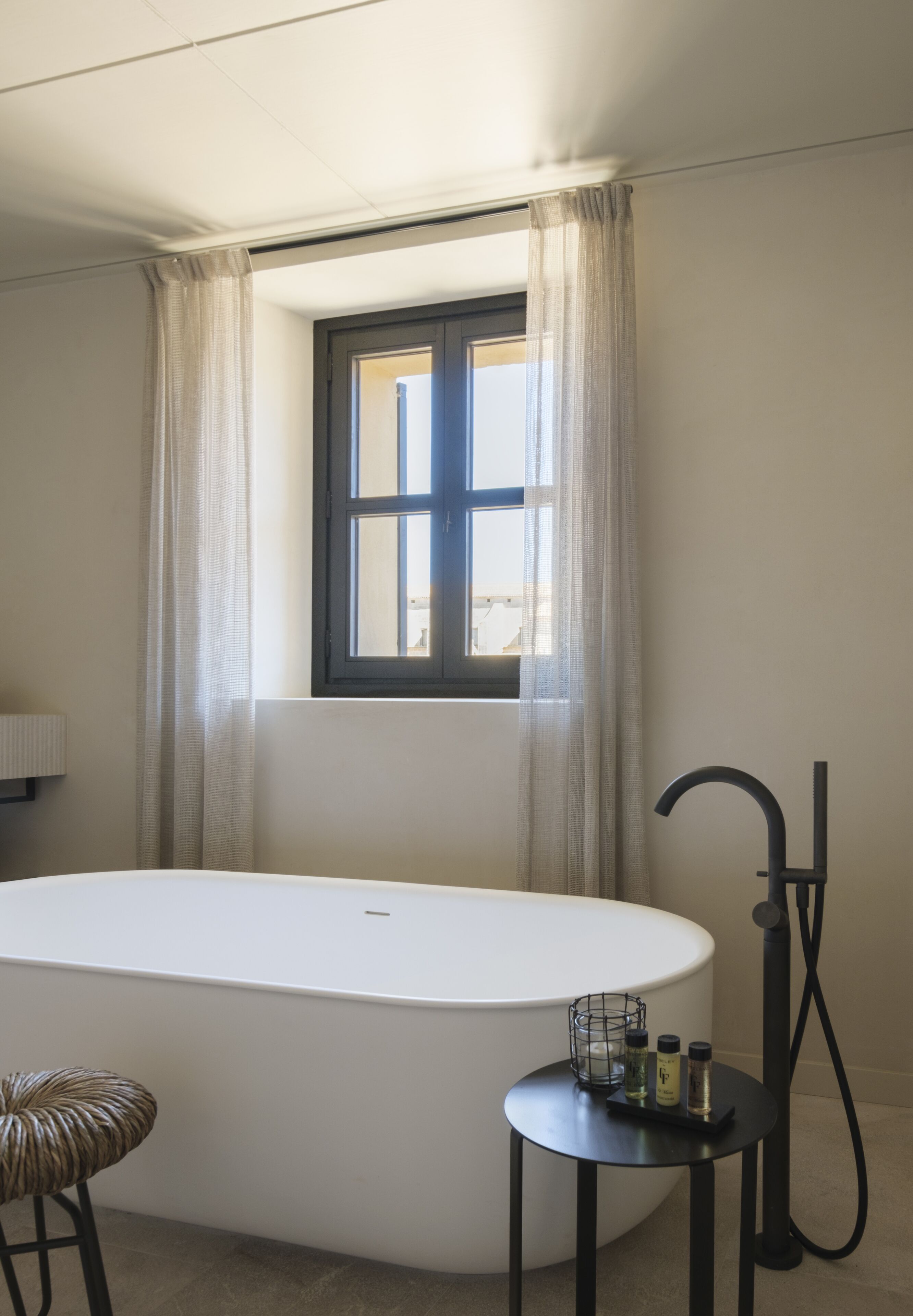 A modern bathroom with a freestanding bathtub, a window with curtains, a towel rack, and a small table with toiletries.