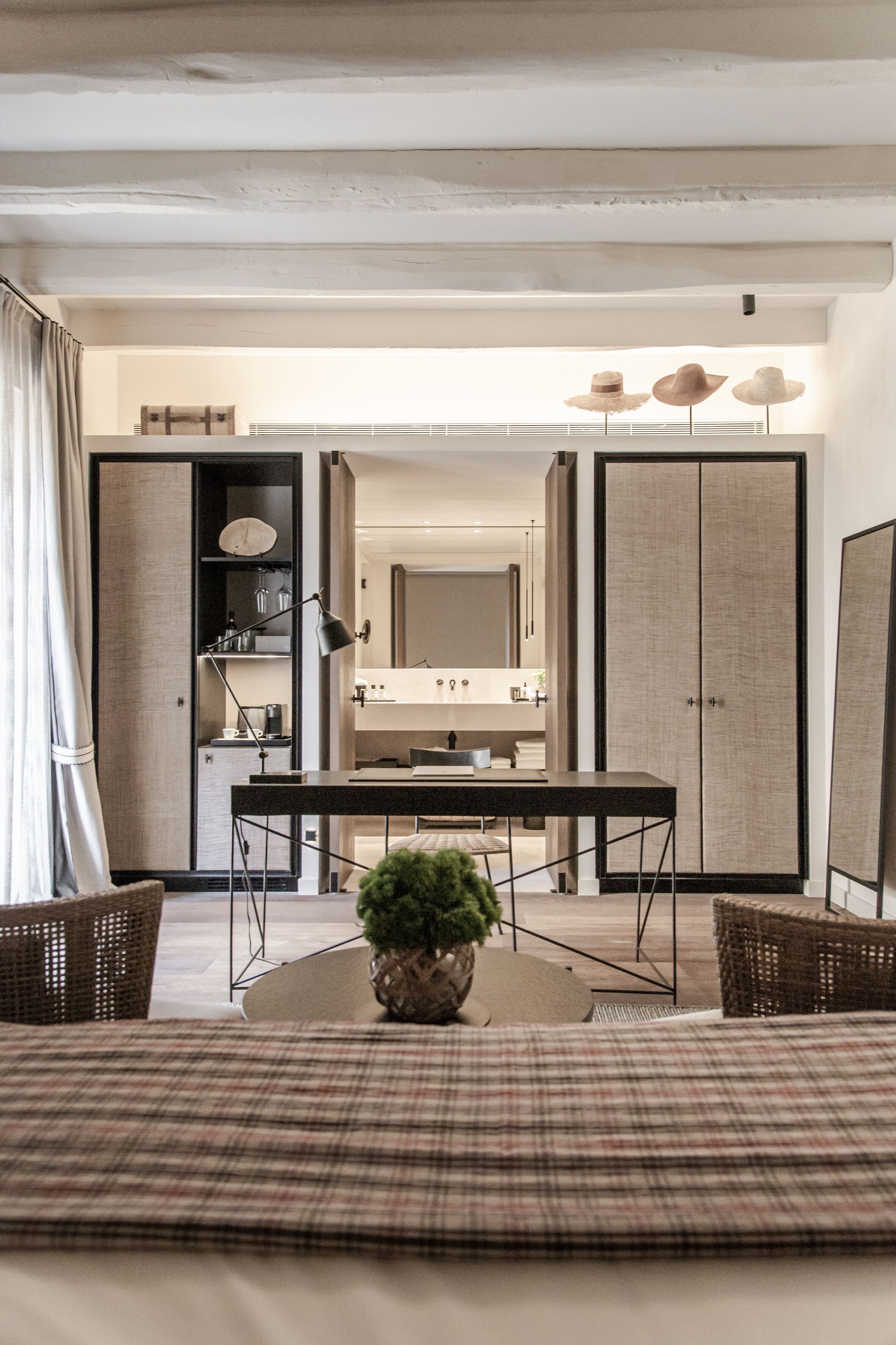 Una habitación de hotel moderna con cama, mesa de comedor, planta en maceta, mobiliario de mimbre y un armario grande.
