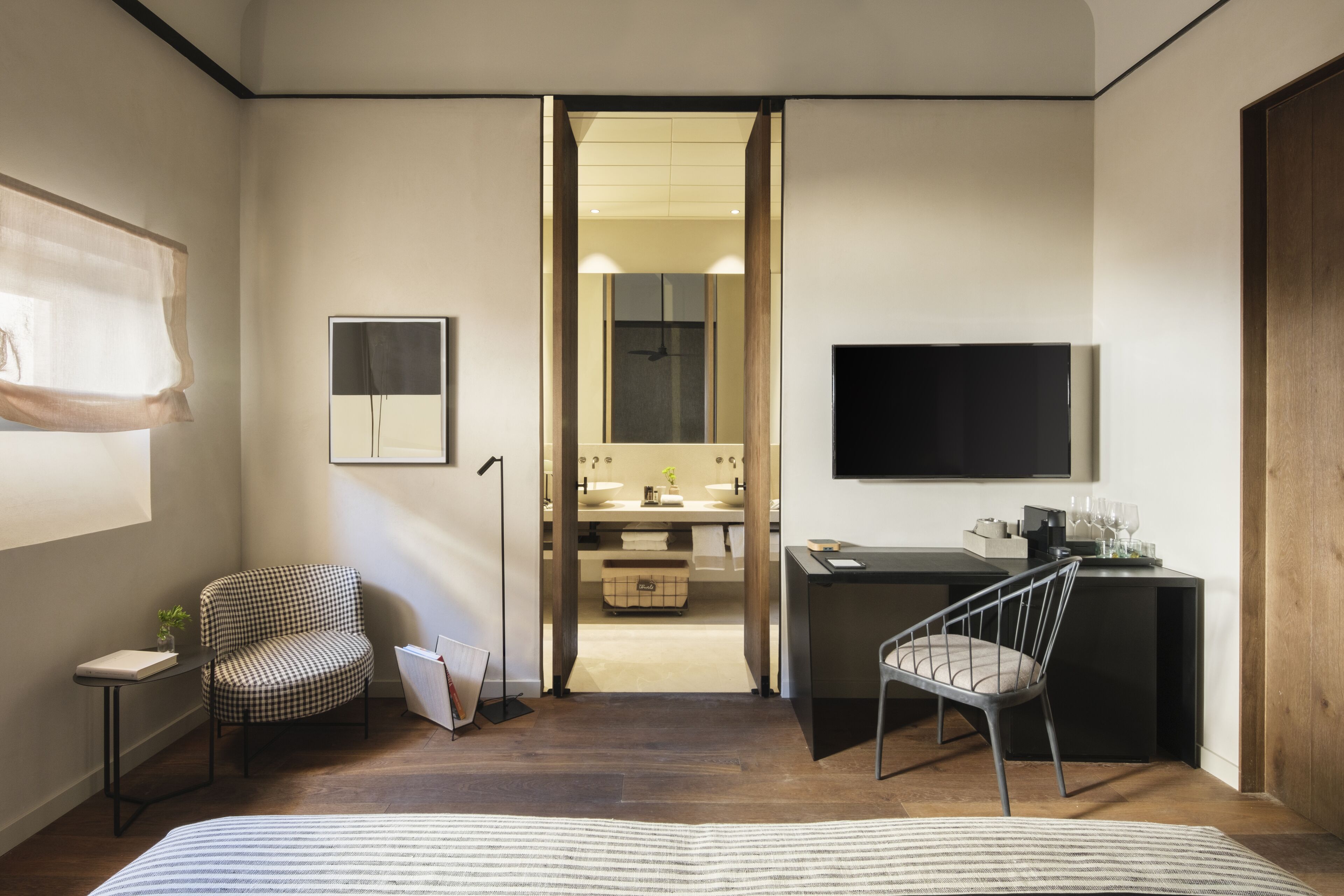 A modern bedroom with a bed, a desk, a chair, a television, and a bathroom visible through an open door.