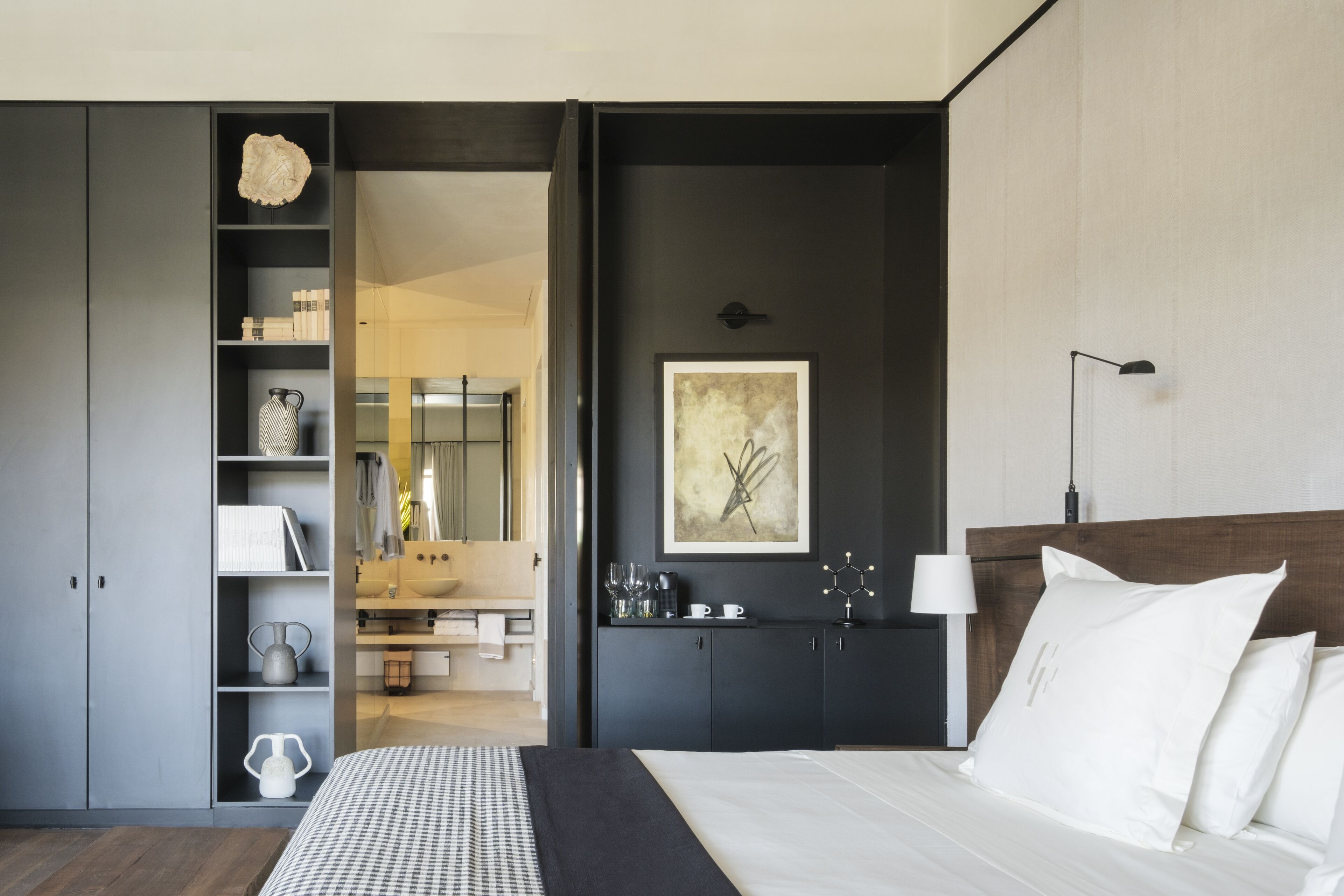 Une chambre moderne avec un grand lit, un mur foncé orné d’un tableau encadré, une table de chevet avec une lampe et une armoire intégrée.