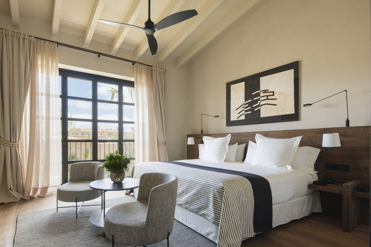 A hotel room with a large bed, two bedside tables, a ceiling fan, a window with curtains, a small table with chairs, and a framed artwork on the wall.