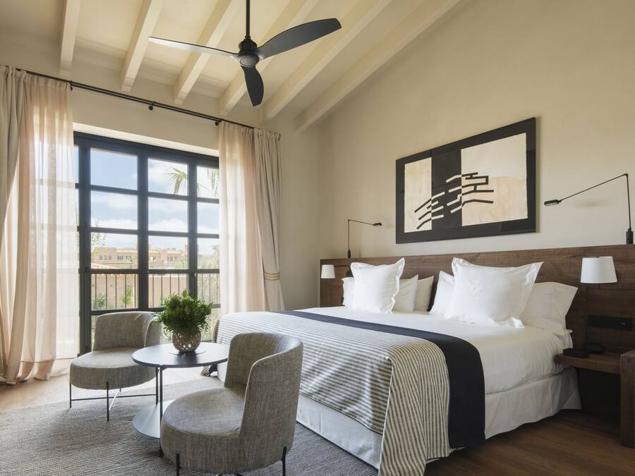 A hotel room with a large bed, two bedside tables, a ceiling fan, a window with curtains, a small table with chairs, and a framed artwork on the wall.