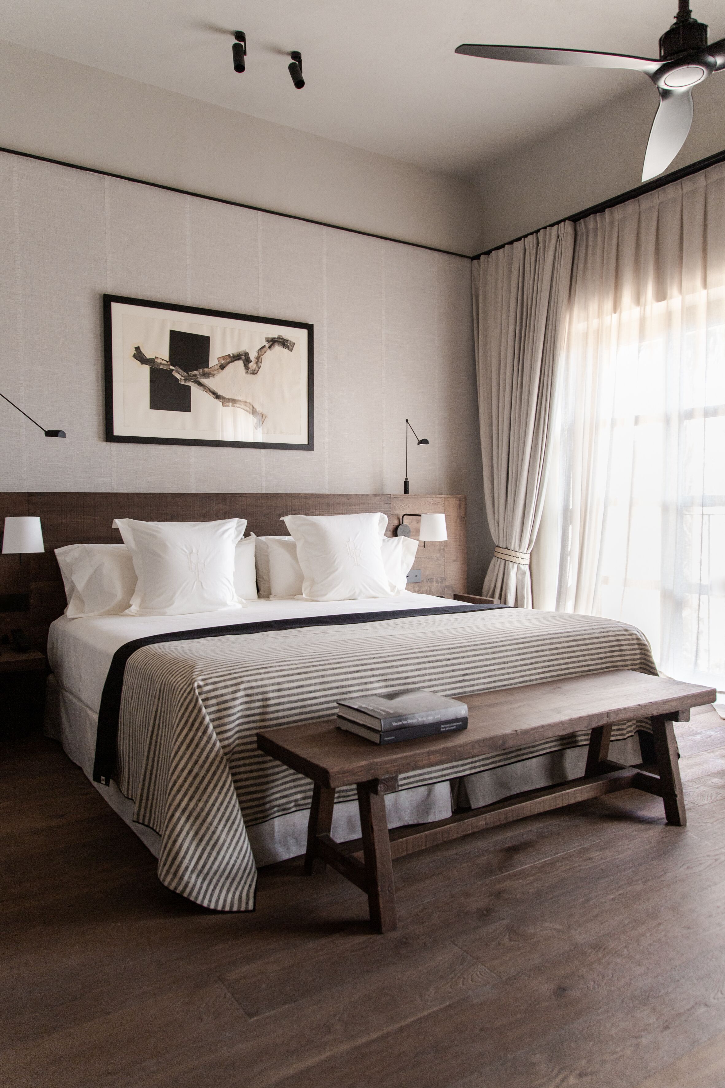 Une chambre d’hôtel moderne avec un grand lit, un banc en bois, un ventilateur de plafond et une œuvre d’art encadrée au mur.