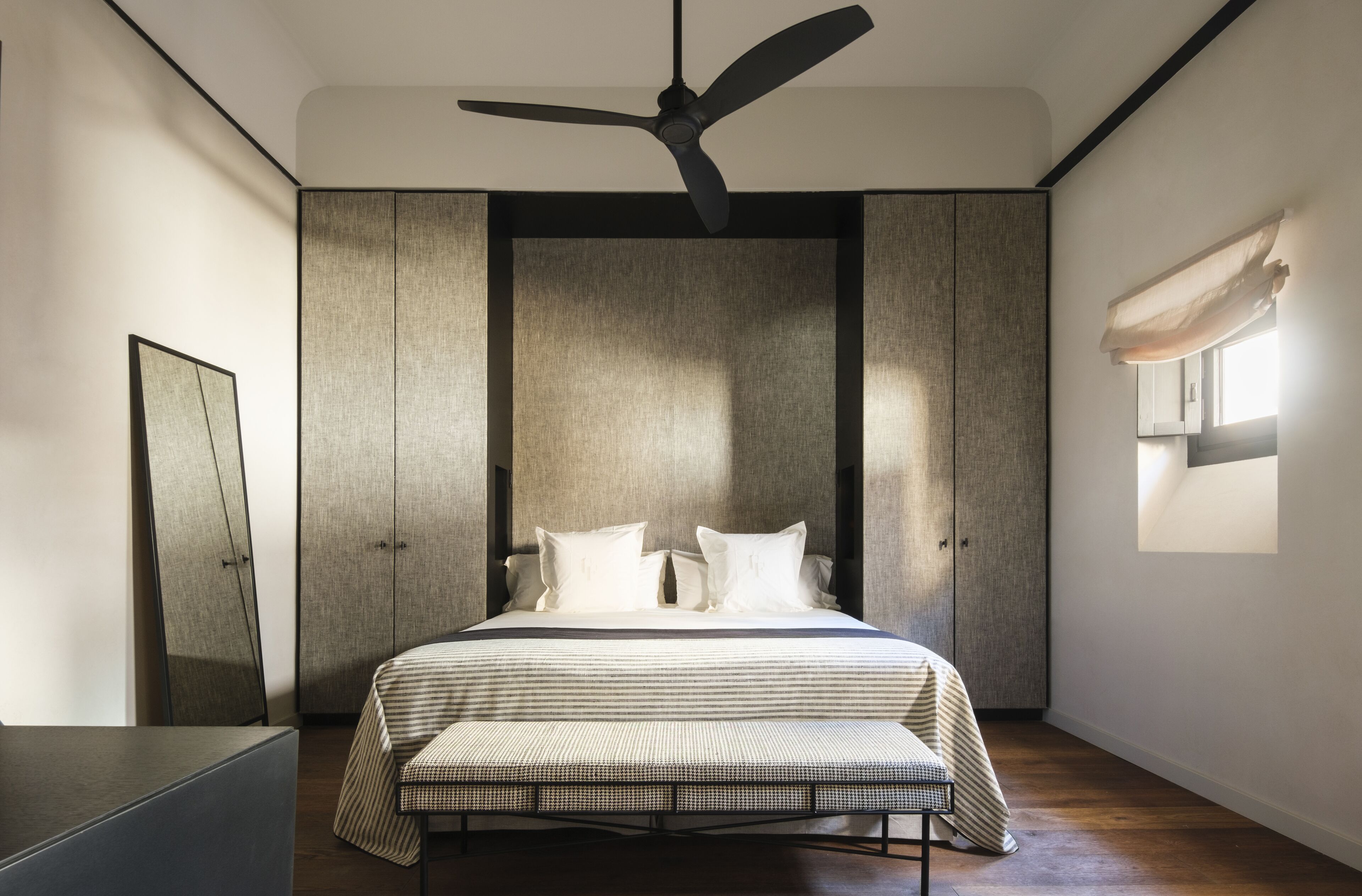 Une chambre moderne avec un lit, un banc, un ventilateur de plafond et une fenêtre avec des rideaux.
