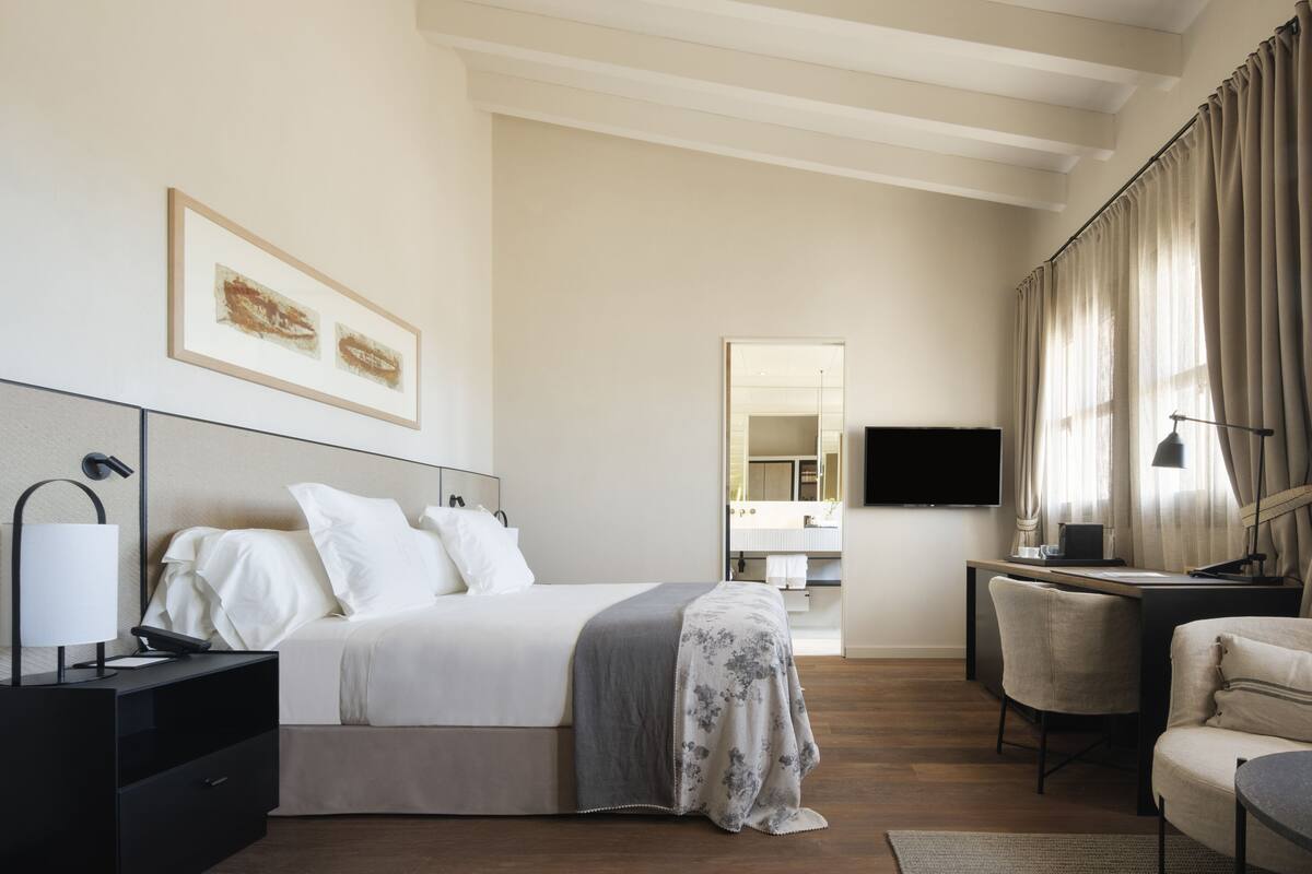 A modern hotel room with a bed, bedside table, lamp, TV, and a desk with a chair.