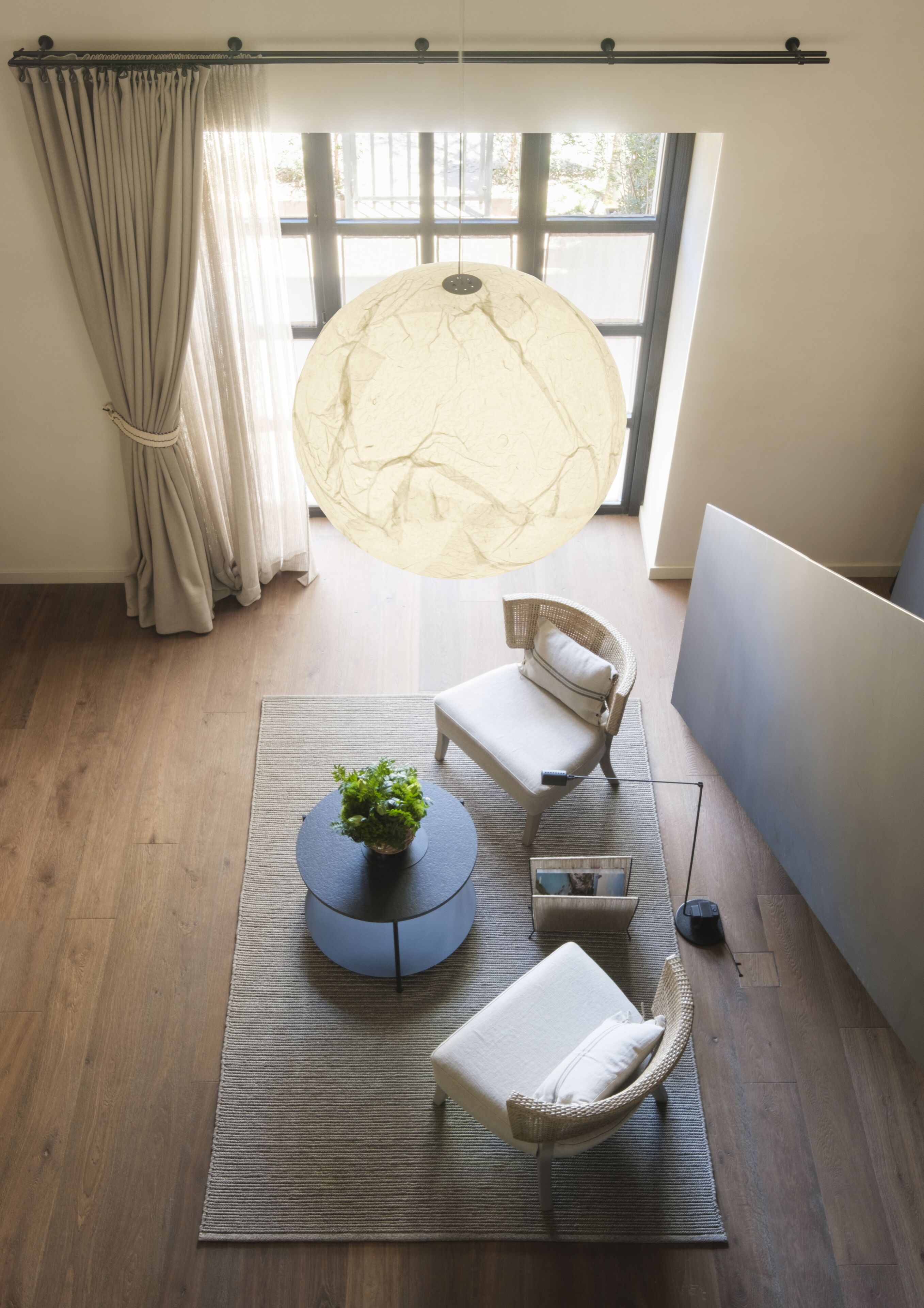 A room with a large window, a round table with a plant, two wicker chairs, and a hanging light fixture.