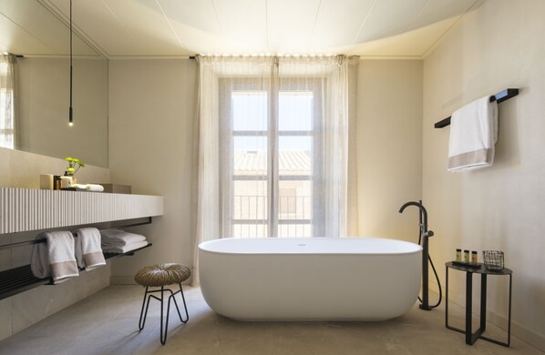 Un baño moderno con una bañera independiente, un espejo grande, un tocador con toallero y una ventana con cortinas de encaje.