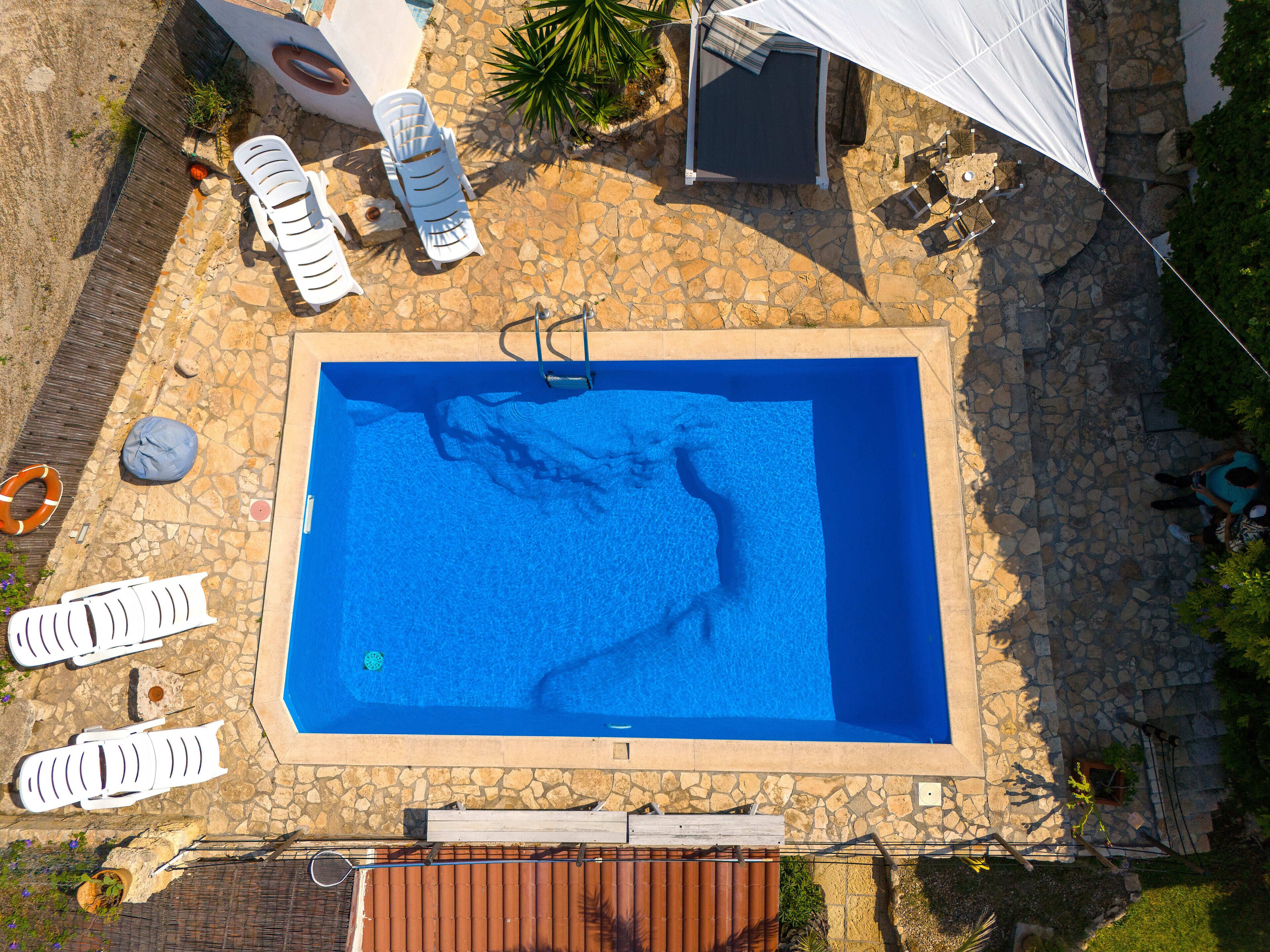 A rectangular pool with a stone border, surrounded by white lounge chairs and a covered area.