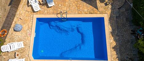 A rectangular pool with a stone border, surrounded by white lounge chairs and a covered area.